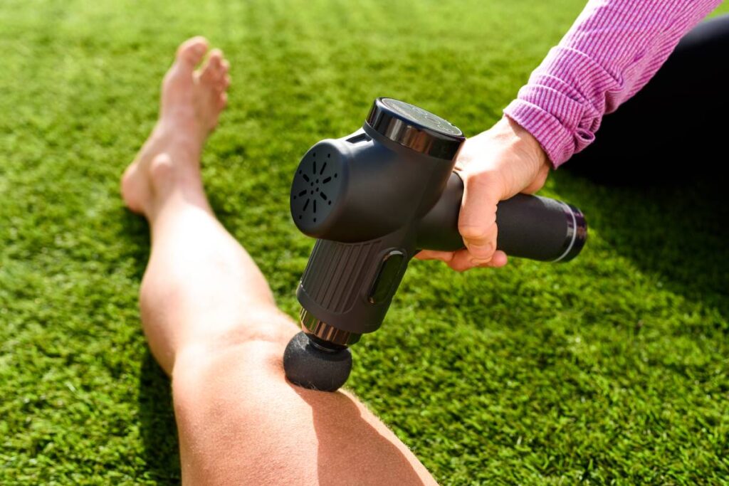 Assistant massages the legs of an athlete with a percussion massage gun to recover from an intense workout.